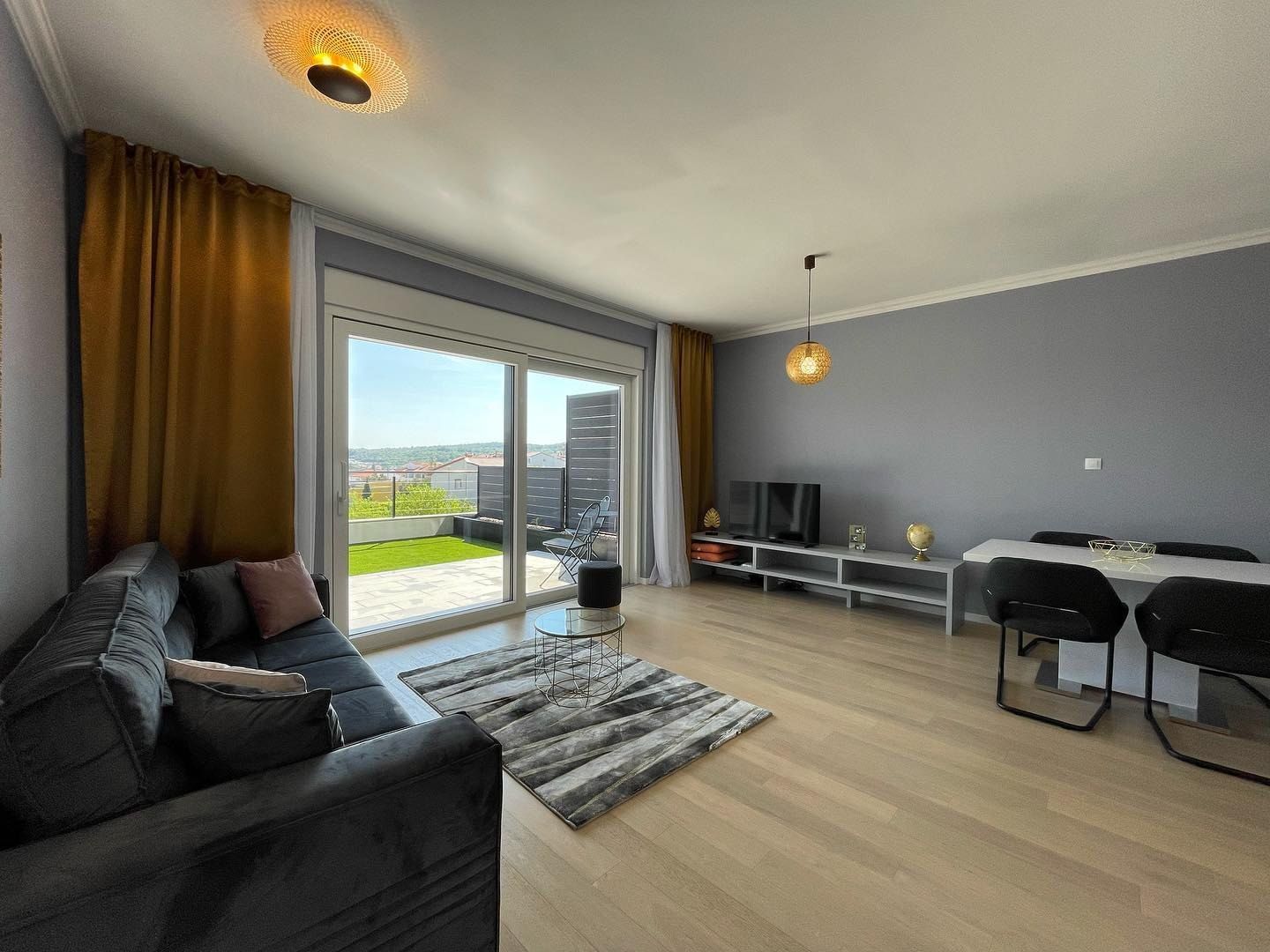 Living room of the Nanea apartment: a grey velvet sofa, a white dining table and glass doors open to a lawn terrace